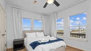 A bedroom with a bed, a nightstand, a ceiling fan, and large windows with a view of the outside.