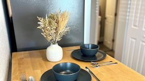 Ein kleiner Holztisch mit einer dunklen Schüssel, einer weißen Vase mit getrockneten Blumen sowie einem Löffel und einer Gabel auf einem Holzboden.