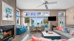 A spacious living room with a fireplace, comfortable seating, a ceiling fan, and large windows overlooking a deck. - Beachview | Fire Pit | Cargo Lift | Pet-Friendly | Steps to Shore (Galveston)