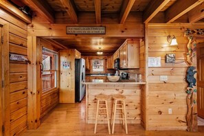 A cabin interior with wooden walls, a kitchen area, and a bar with stools. - Smoky Mountain Views | Hot Tub | Game Room | Sleeps 12 (Sevierville)