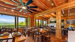 A wooden interior with dining tables, chairs, a ceiling fan, and large windows offering a view of the outdoors. - Luxury 12BR Lodge Sleeps 60 | Heated Pool + Pickleball Court + Sauna + Putt Putt (Sevierville)