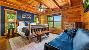 A log cabin bedroom with a bed, a bench, a sofa, a ceiling fan, and large windows. - Luxury 12BR Lodge Sleeps 60 | Heated Pool + Pickleball Court + Sauna + Putt Putt (Sevierville)
