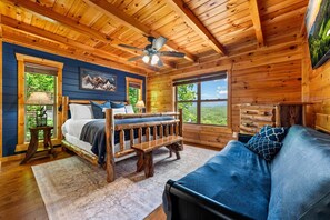Ein Schlafzimmer in einer Blockhütte mit einem Bett, einer Bank, einem Sofa, einem Deckenventilator und großen Fenstern.