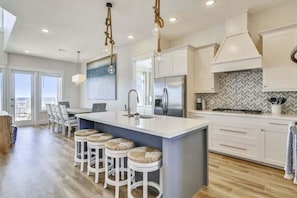 A modern kitchen with a central island, bar stools, a large refrigerator, and a herringbone-patterned backsplash. - Luxury 5BR Beachfront Home | Sleeps 14 | Community Pool (Galveston)