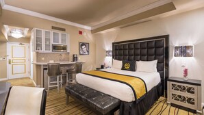 Habitación de hotel con una cama grande, un escritorio con silla, una kitchenette y una mesita de noche con lámpara.