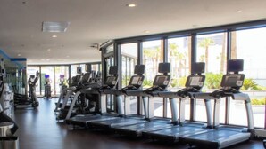 Un gimnasio con cintas de correr y elípticas, ventanas amplias y vista a palmeras desde el exterior.