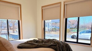 Ein Schlafzimmer mit einem Bett, einem Nachttisch mit Lampe und großen Fenstern mit Jalousien.