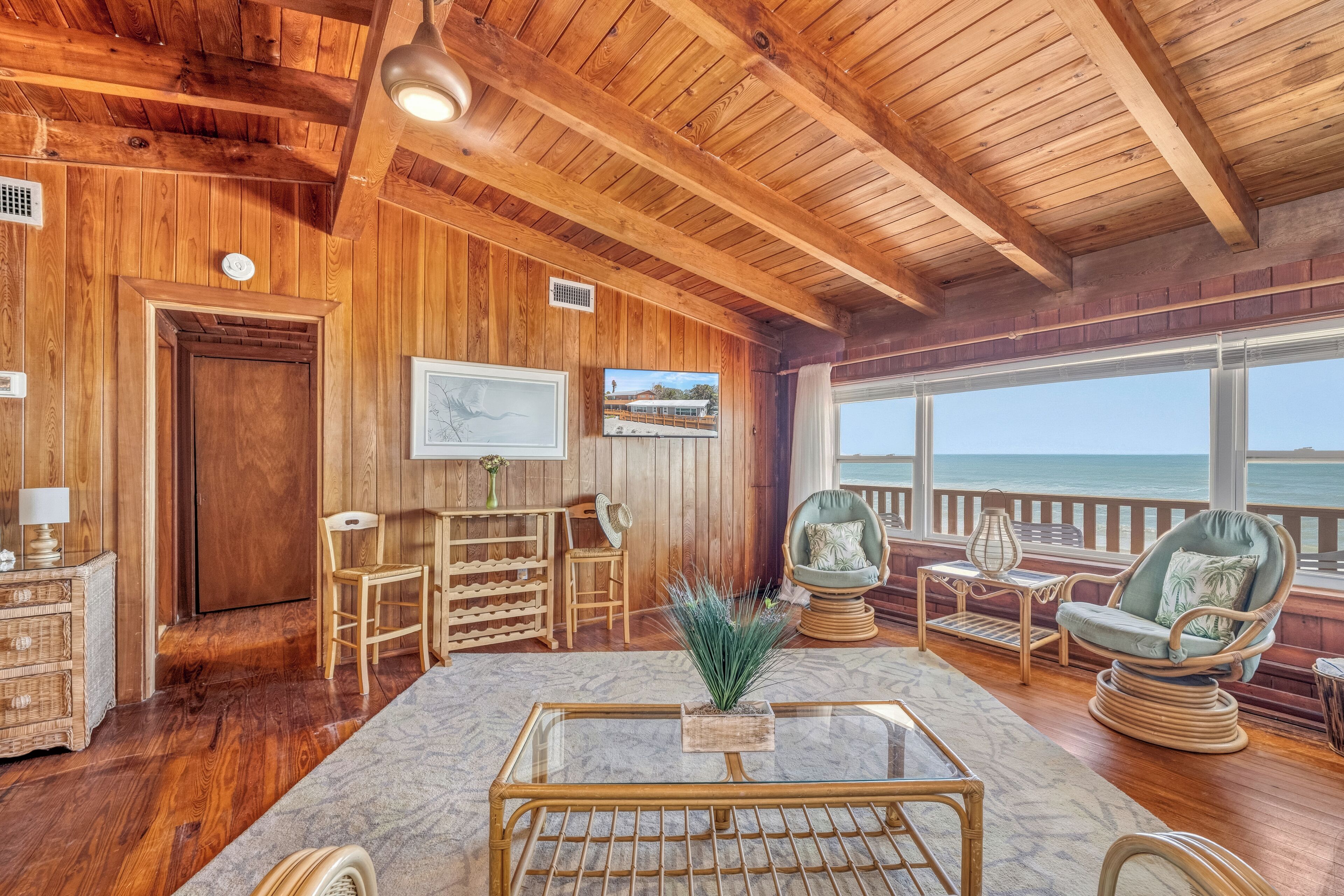 Ein Holzinterieur mit einem Glastisch, Korbstühlen und Blick aufs Meer.