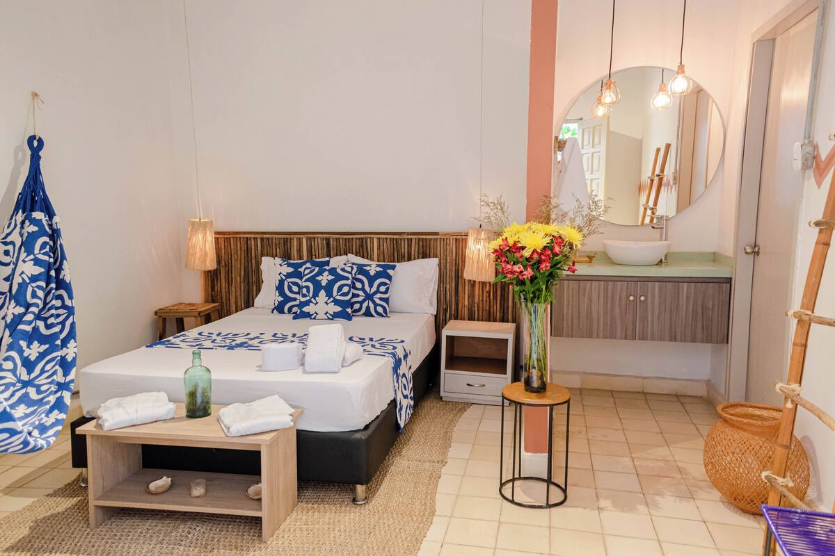 Un dormitorio con cama, mesitas de noche, tocador, espejo, baño con lavabo y un florero con flores.