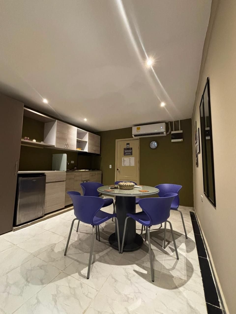 Una cocina compacta con electrodomésticos modernos, un comedor con una mesa redonda y cuatro sillas, y un equipo de aire acondicionado de pared.