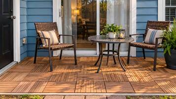 Une terrasse en bois avec deux fauteuils en osier et une table, une plante en pot et une porte donnant sur un espace intérieur.