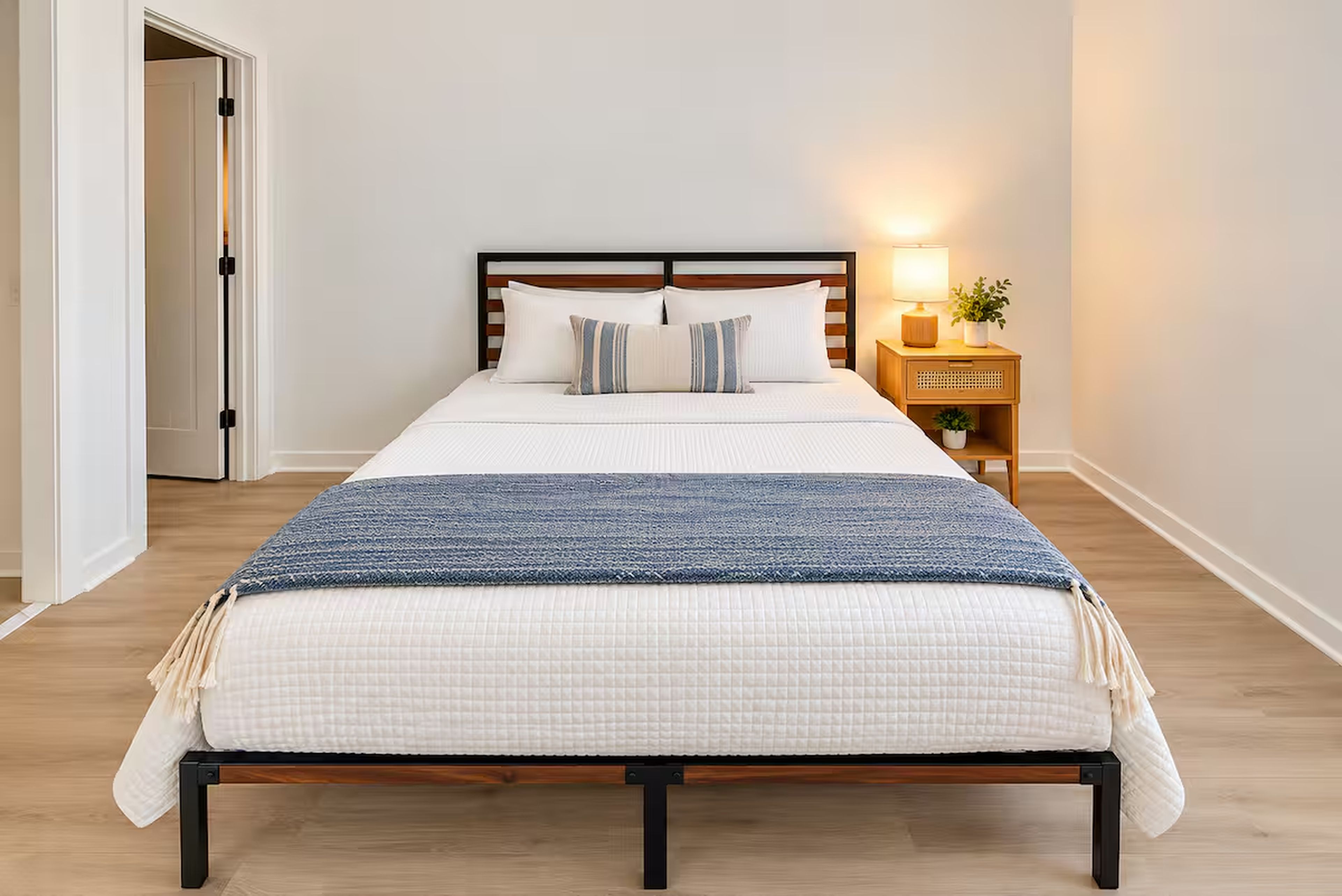 Cama com estrutura de madeira, roupa de cama branca e manta azul. Há uma mesa de cabeceira com abajur e uma planta.