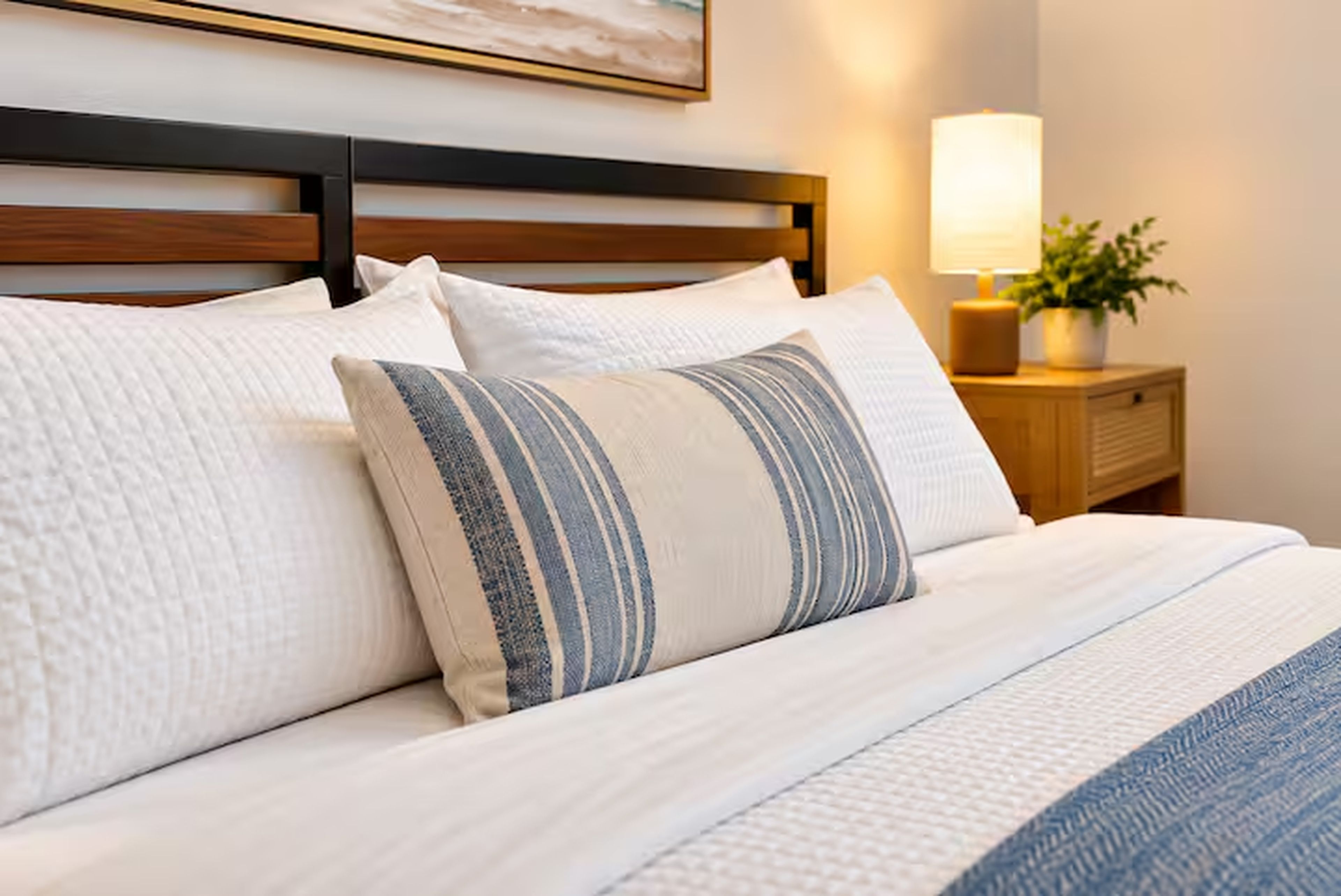 A neatly made bed with a white duvet and striped pillows, a wooden headboard, a bedside lamp, and a small plant on a wooden nightstand.