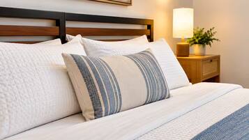A neatly made bed with a white duvet and striped pillows, a wooden headboard, a bedside lamp, and a small plant on a wooden nightstand.
