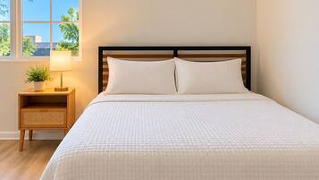 A neatly made bed with a quilted white duvet, a wooden headboard, and a bedside table with a lamp and a small plant.