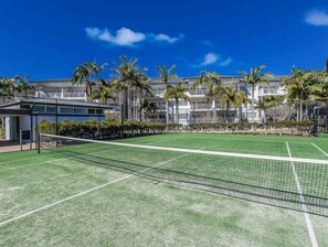 Een tennisbaan met een net, omringd door palmbomen en een gebouw met balkons.
