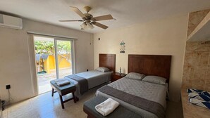 A hotel room with two beds, a ceiling fan, a bench, a small table, and a view of the outside.