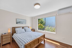 Ein Schlafzimmer mit einem hölzernen Bett, einem Nachttisch, einer Nachttischlampe, einem Fenster mit Blick ins Grüne und einer Deckenleuchte.