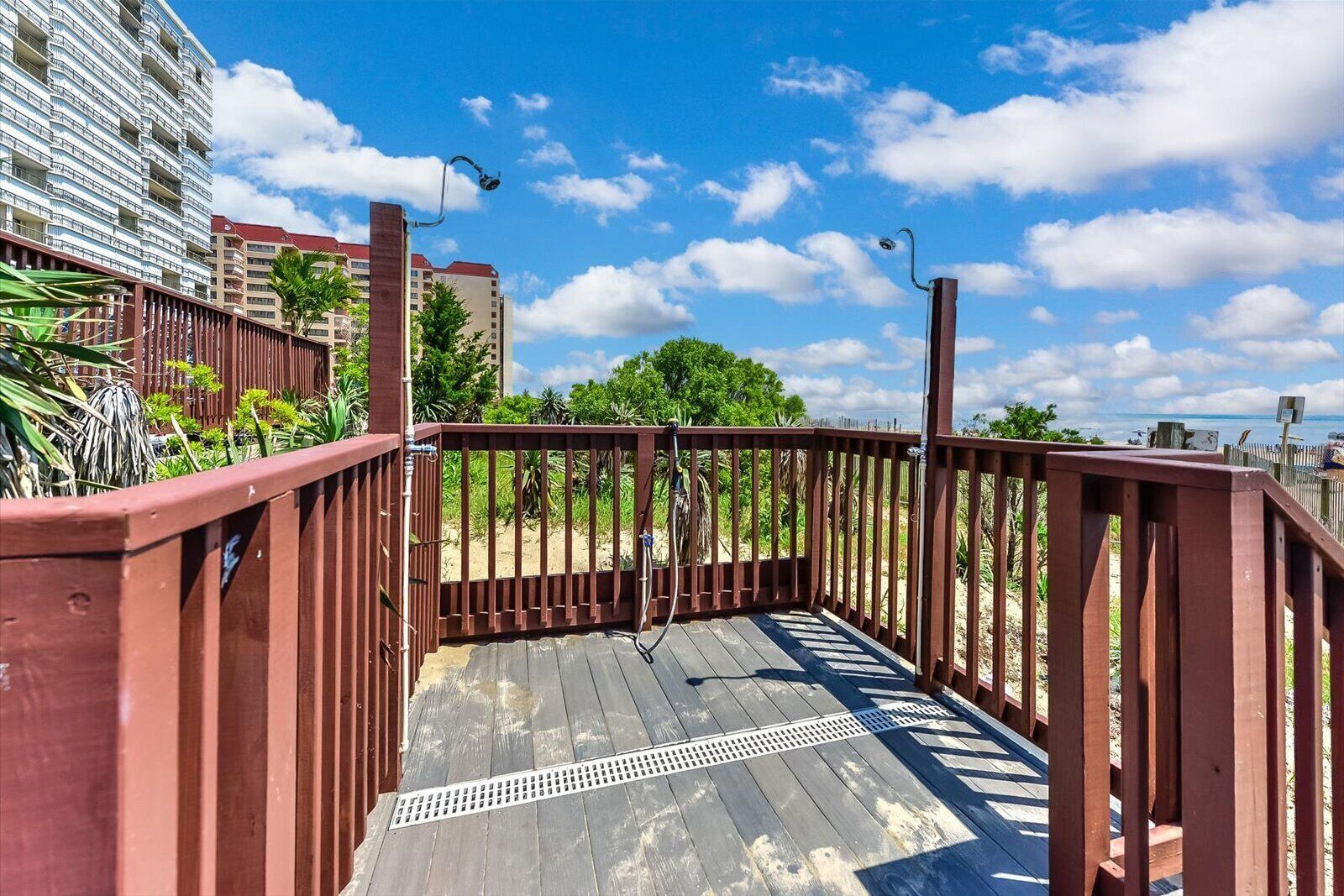 Une terrasse en bois avec des garde-corps, une grille métallique et une vue sur une plage et des bâtiments.