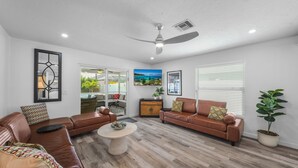 A living room with two brown leather sofas, a round coffee table, a ceiling fan, and a large window with blinds. - New! Gulf Breeze - Stunning Heated Saltwater Pool & Spa with Sundeck, Apprx 1 Mile to Beach! (Naples)