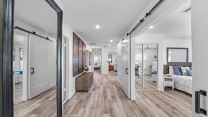 A hallway with wooden flooring, white walls, and a bed with a gray headboard and blue pillows. - New! Gulf Breeze - Stunning Heated Saltwater Pool & Spa with Sundeck, Apprx 1 Mile to Beach! (Naples)
