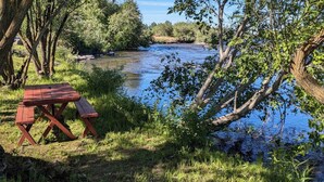 Piknikpöytä joen varrella, ympärillä puita ja vehreää kasvillisuutta.