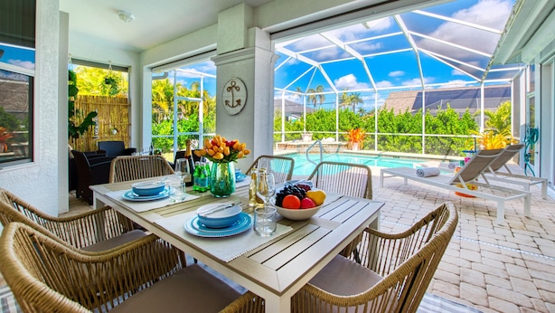 Un patio techado con una mesa de comedor preparada para cuatro personas, rodeado de sillas de mimbre, con vista a una piscina y abundante vegetación.