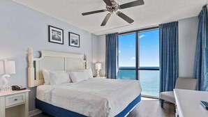 Ein Hotelzimmer mit Bett, Schreibtisch, Stuhl, Deckenventilator und einem großen Fenster mit Vorhängen.