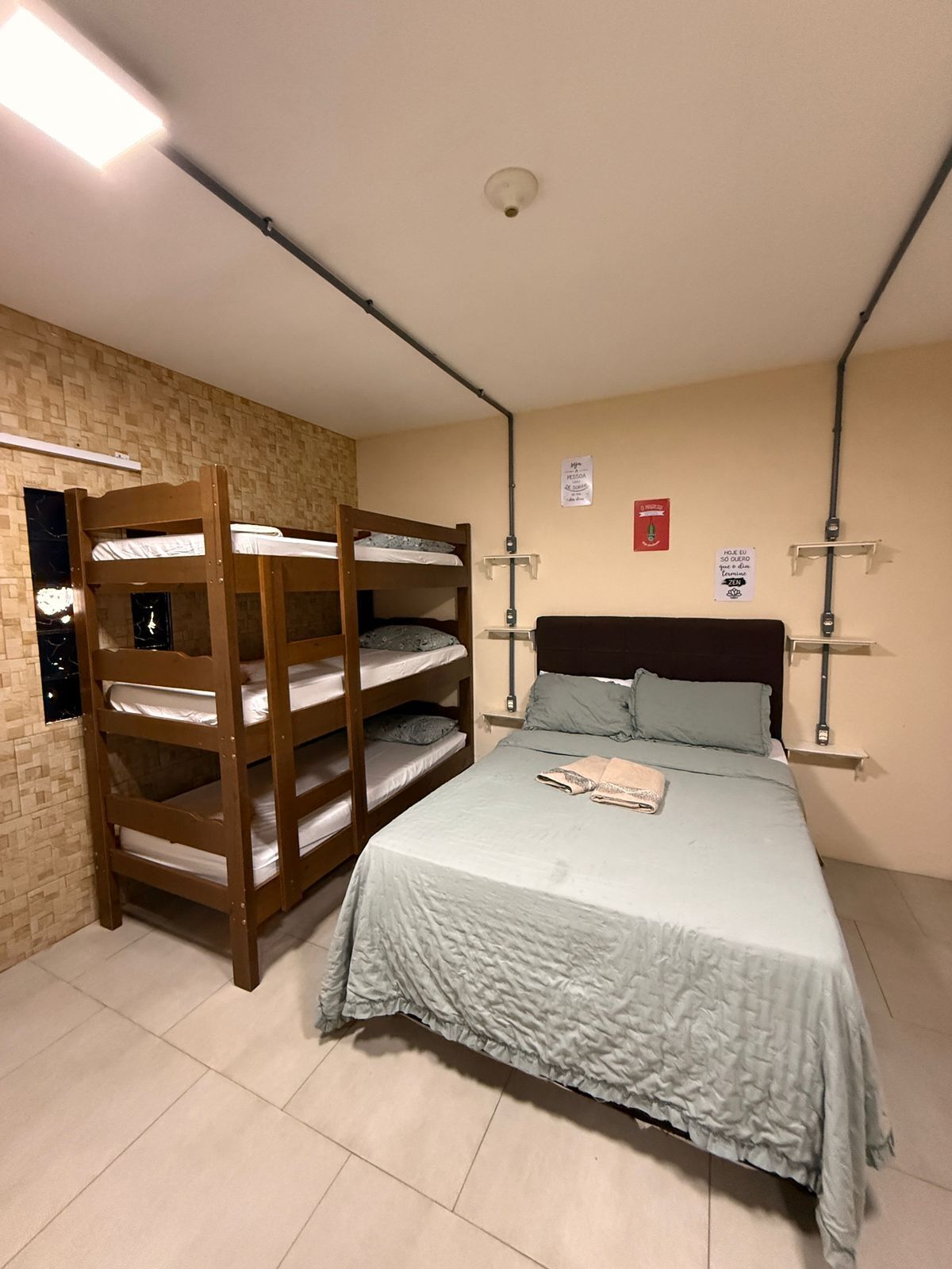 A bedroom with a bunk bed, a single bed, a brick wall, a ceiling light, and a fire alarm.