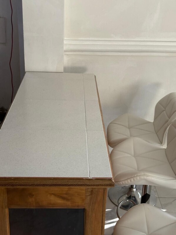 A wooden table with a white top, positioned next to a white wall and a chair with a cushioned seat.