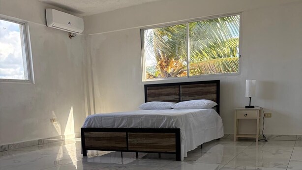 A bedroom with a bed, a nightstand, a window with a view of palm trees, and an air conditioning unit.