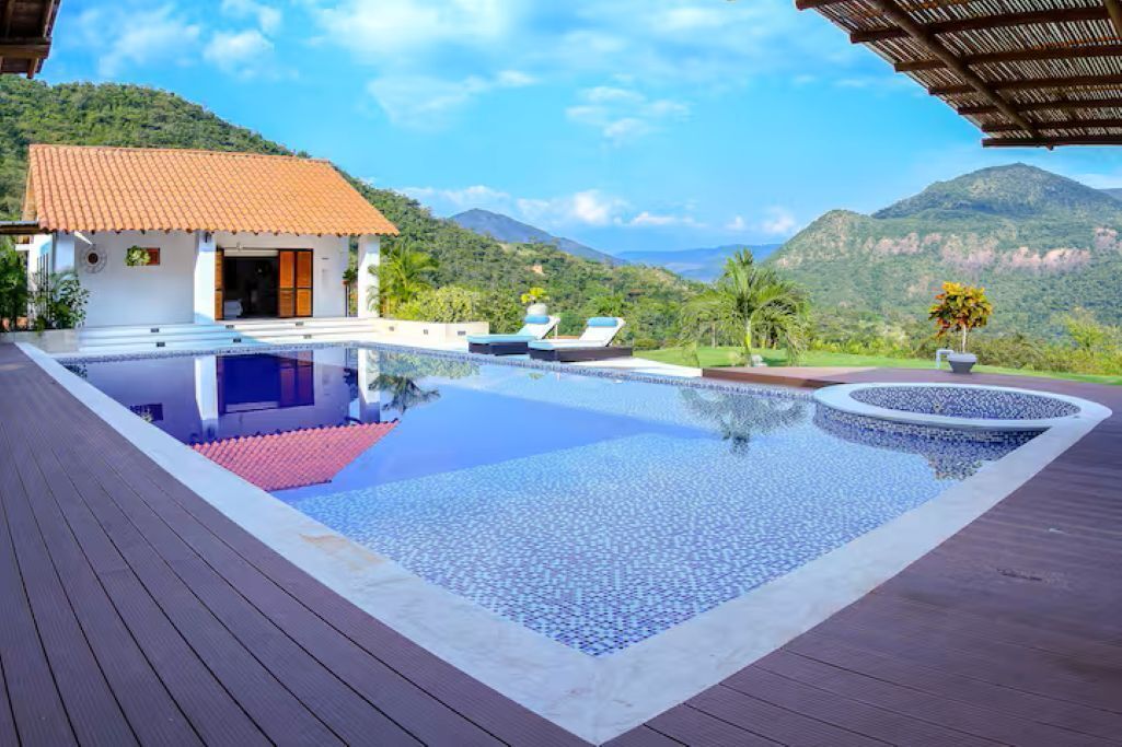 A pool with a wooden deck, a small building with a red roof, and a mountainous backdrop.