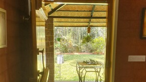 Une porte-fenêtre donnant sur un patio équipé d’une table et de chaises, d’un paillasson et offrant une vue sur un jardin.