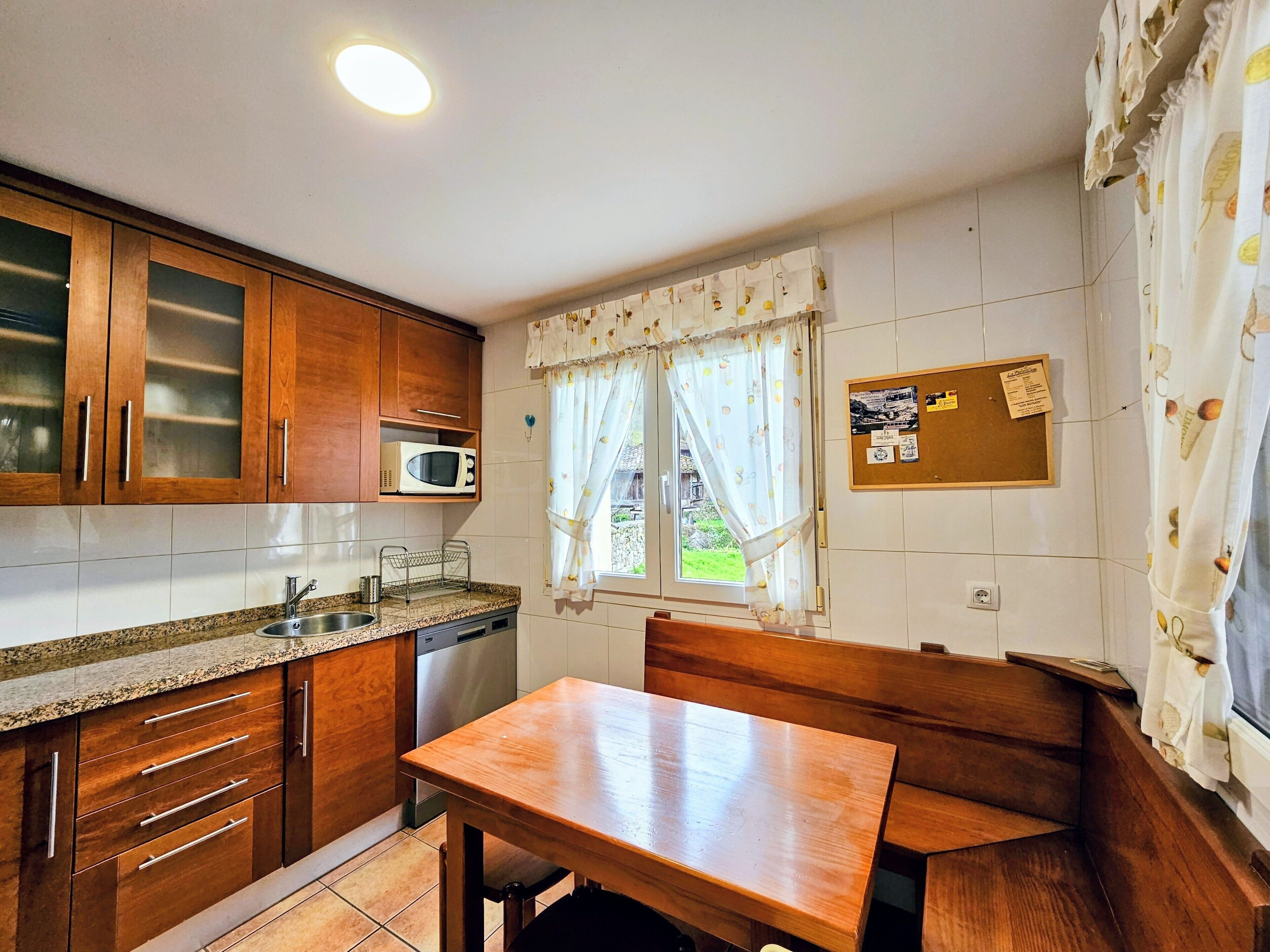 Een compacte keuken met houten kastjes, een magnetron, een spoelbak en een eettafel met bankjes.