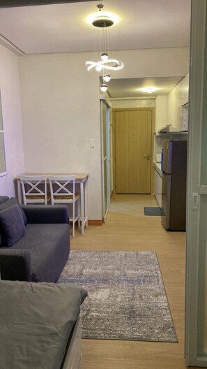 A compact living space with a sofa, a small table, and a kitchen area with a refrigerator. - Cozy Corner Staycation | Trees Residences (Quezon City)