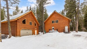Eine Blockhütte mit zwei angebauten Garagen, umgeben von Schnee und hohen Kiefern.