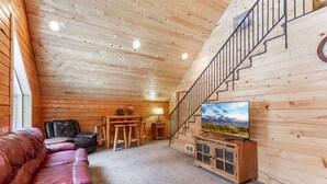 Ein gemütliches Blockhaushaus-Interieur mit Holzbalkendecke, einer Treppe, einem Ledersofa, einem Holz-TV-Möbel und Blick auf die Berge.