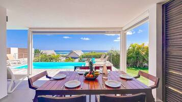 Un coin repas avec une table dressée pour quatre personnes, donnant sur une piscine et un paysage tropical.
