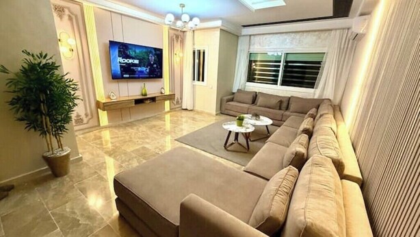 A modern living room with a large sectional sofa, a flat-screen TV mounted on the wall, and a potted plant. - Reda's'Apparts (Downtown - Wifi 200 Mb) (Fès)