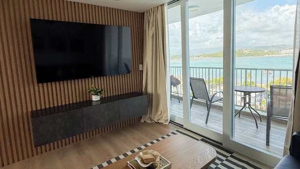 A modern living room with a flat-screen TV, wooden panel wall, and a balcony with a view of the sea. - The Oasis - Private Island Getaway! (Fajardo)