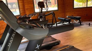 Un gimnasio con varios equipos de ejercicio, incluyendo cintas de correr y una máquina elíptica, en una sala con paredes de madera y ventanas amplias.