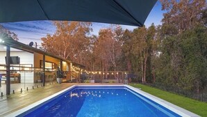 An outdoor swimming pool with a wooden deck, surrounded by trees and a covered area with string lights.