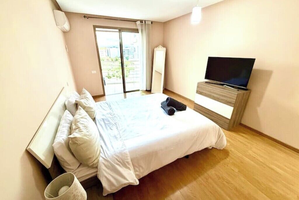 Ein Schlafzimmer mit einem Bett, einem Fernseher auf einem Schrank und Blick ins Freie durch eine Schiebetür.