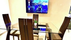 A modern dining area with a glass table and dark wooden chairs. A flat-screen TV mounted on the wall displays a program. - Apartment with park view (Wifi Fiber 100 Mb) (Fès)