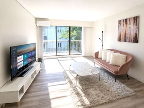 A modern living room with a flat-screen TV, a sofa, a coffee table, and a large window with a view of a balcony. - Lower Lonsdale One Bedroom Apartment - Mins to Seabus/Shipyard (North Vancouver)