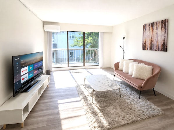 Ein modernes Wohnzimmer mit Fernseher, einer Couch, einem Couchtisch und einem großen Fenster mit Blick auf einen Balkon.