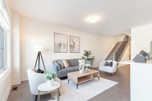 Una sala moderna con un sofá gris, un sillón blanco, una mesita de centro y una lámpara de pie. Se ve una escalera que sube a otro nivel.