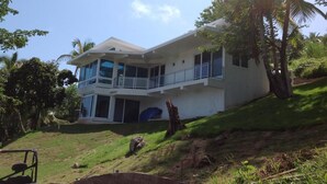 A two-story house with a balcony, surrounded by greenery and palm trees.
