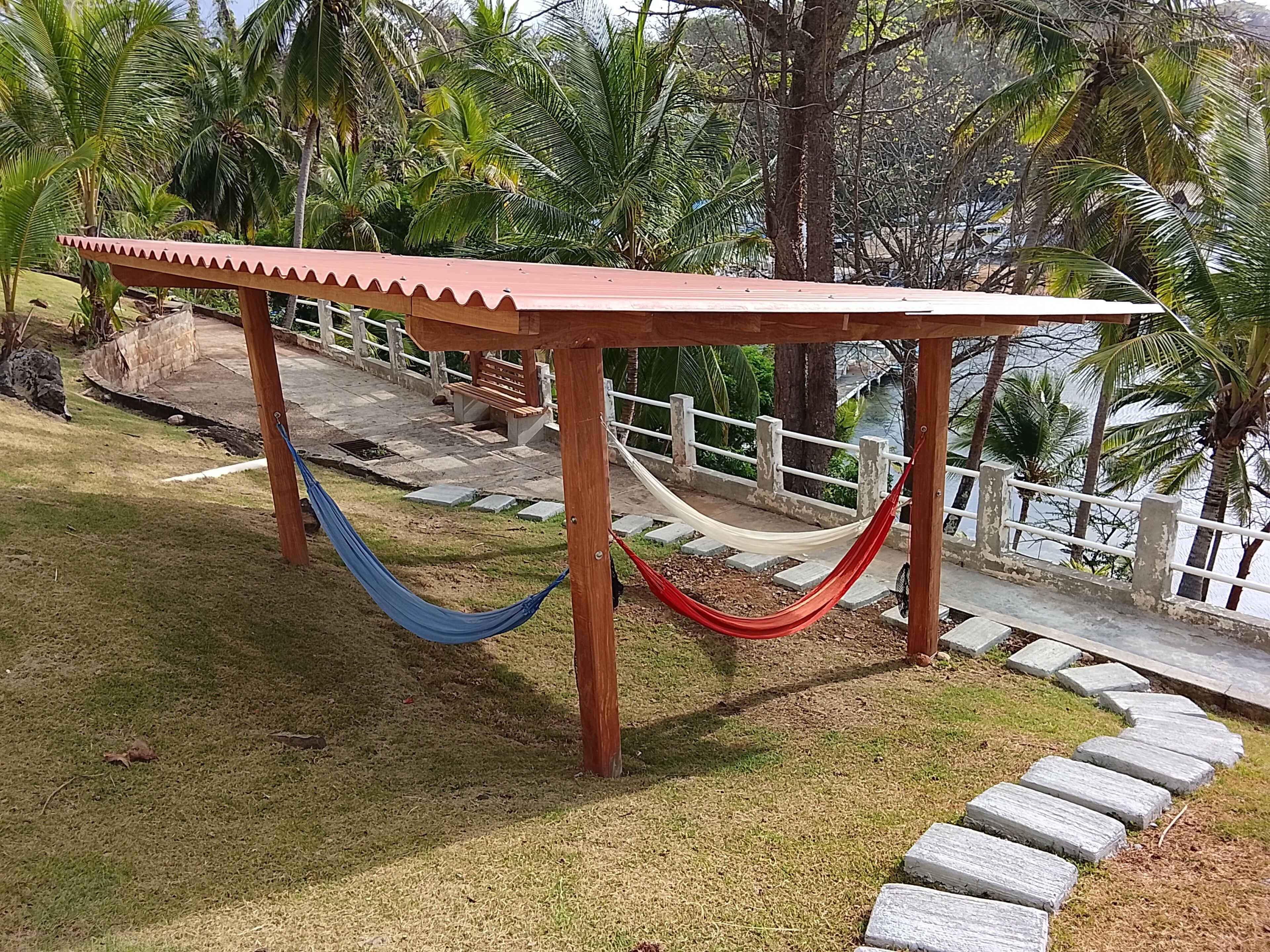 Un hamac sous une pergola en bois, avec un chemin de pierre et des palmiers en arrière-plan.