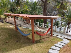 Un hamac sous une pergola en bois, avec un chemin de pierre et des palmiers en arrière-plan.